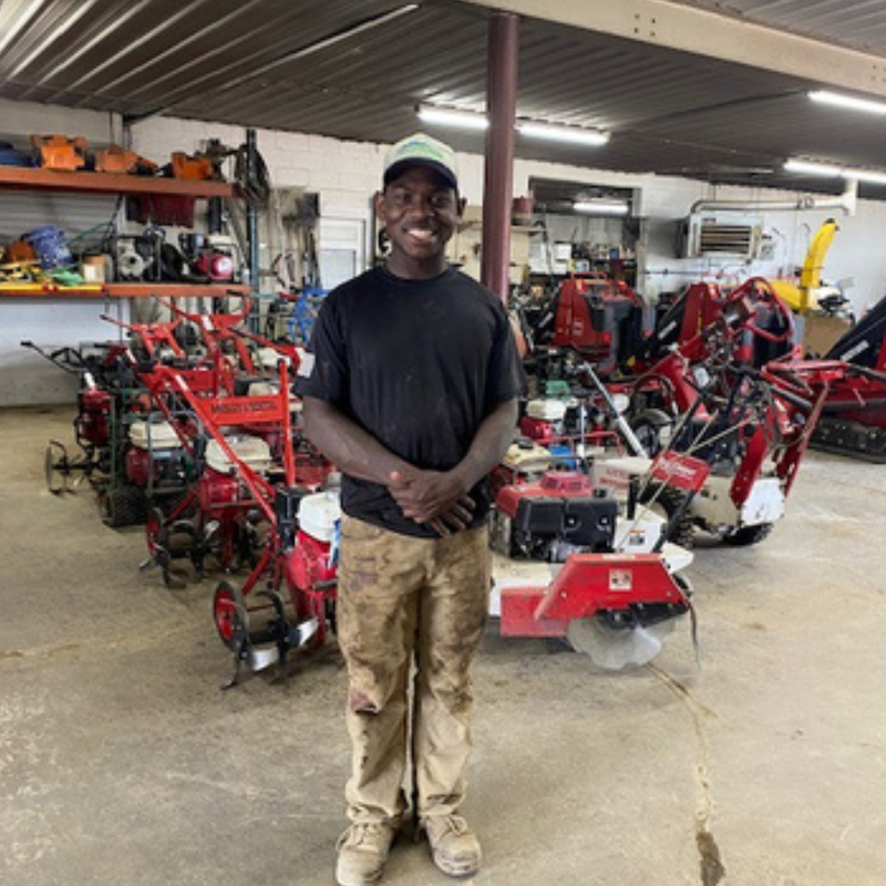 Robbie standing in front of lawnmowers wearing a black shirt and brown pants.